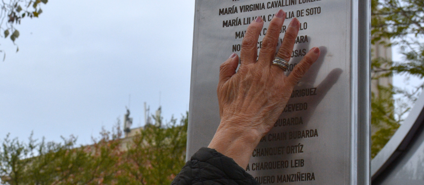 Fotografía. Una mujer pasa su mano en una estructura de metal dónde están los nombres de mujeres víctimas del terrorismo de Estado.